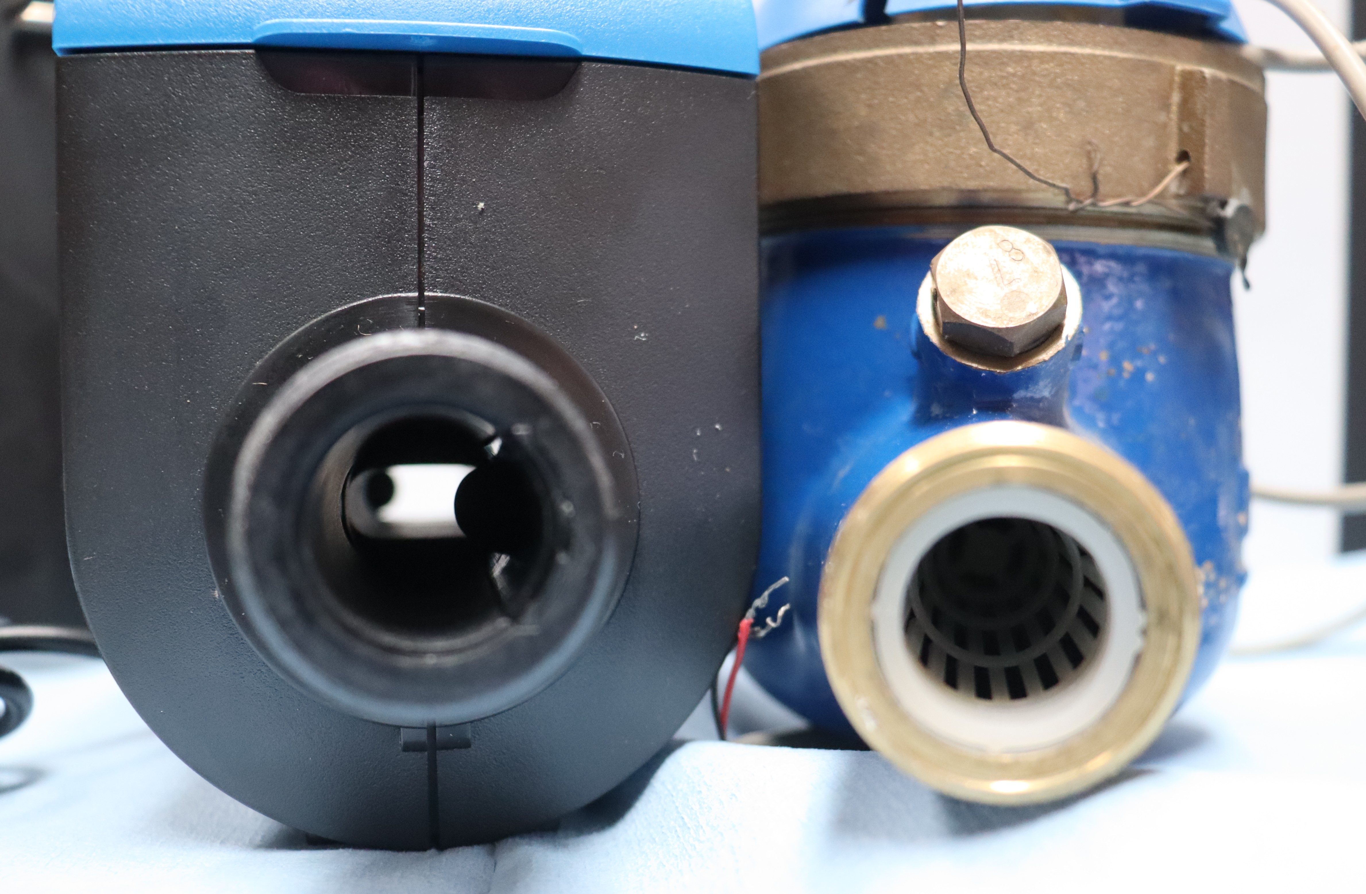 A close-up image of two water meters side by side, showing the front-facing connection points. On the left is a modern black and blue water meter, while on the right is an older, blue metal meter with brass components. Both meters have open inlet or outlet ports, with visible internal structures for water flow. Wires are attached to the older meter, suggesting additional functionality or repairs.